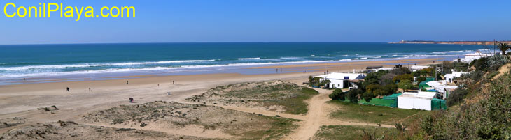 Vista panorámica de la playa con casas blancas y dunas bajo cielo azul despejado y mar tranquilo.
