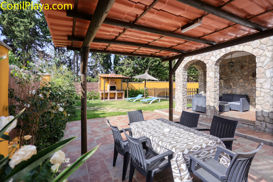 Porche techado con mesa y sillas junto a jardín con barbacoa y tumbonas