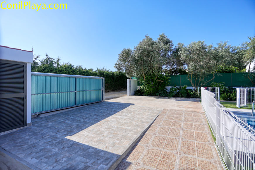 Aparcamiento exterior pavimentado junto a piscina y portón metálico corredero