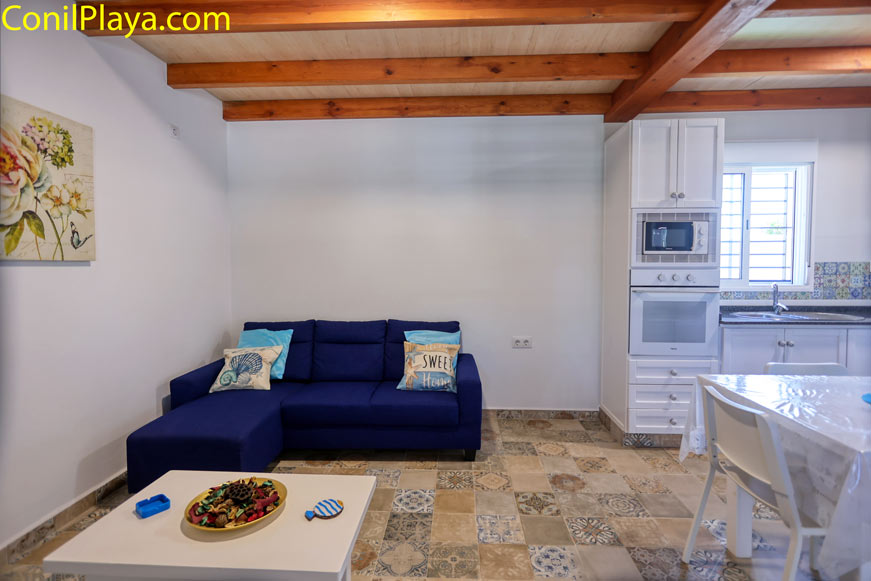 Salón con sofá azul, mesa blanca y cocina integrada visible