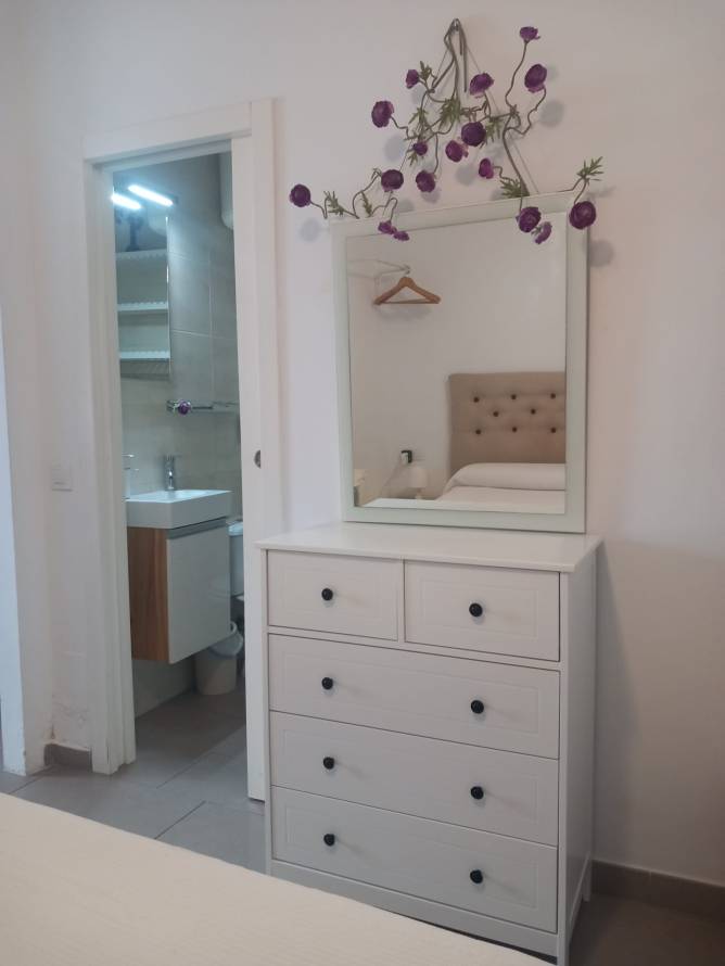 Cómoda blanca con espejo y flores cerca de cama y baño moderno