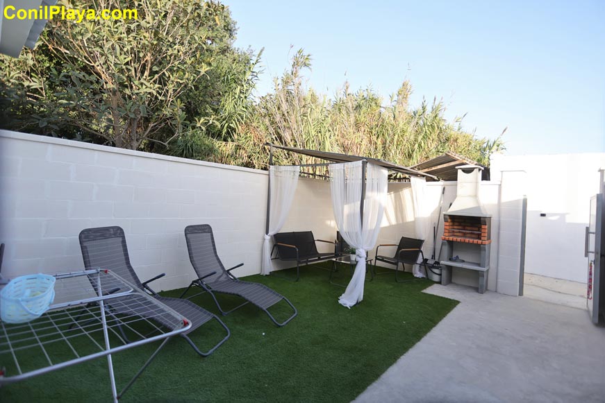 Jardín con tumbonas, pérgola con cortinas y zona de barbacoa junto a pared blanca