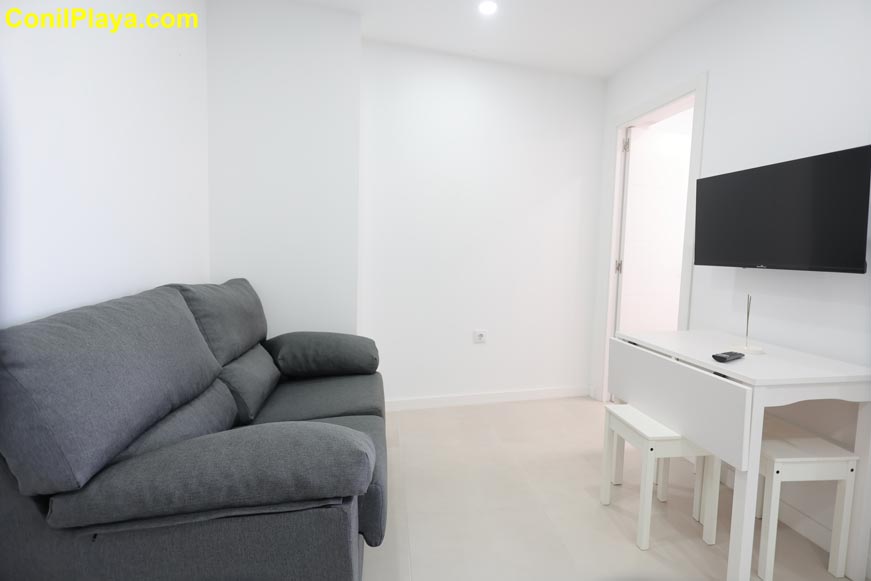 Vista del salón minimalista con sofá gris y televisión en la pared junto a mesa blanca