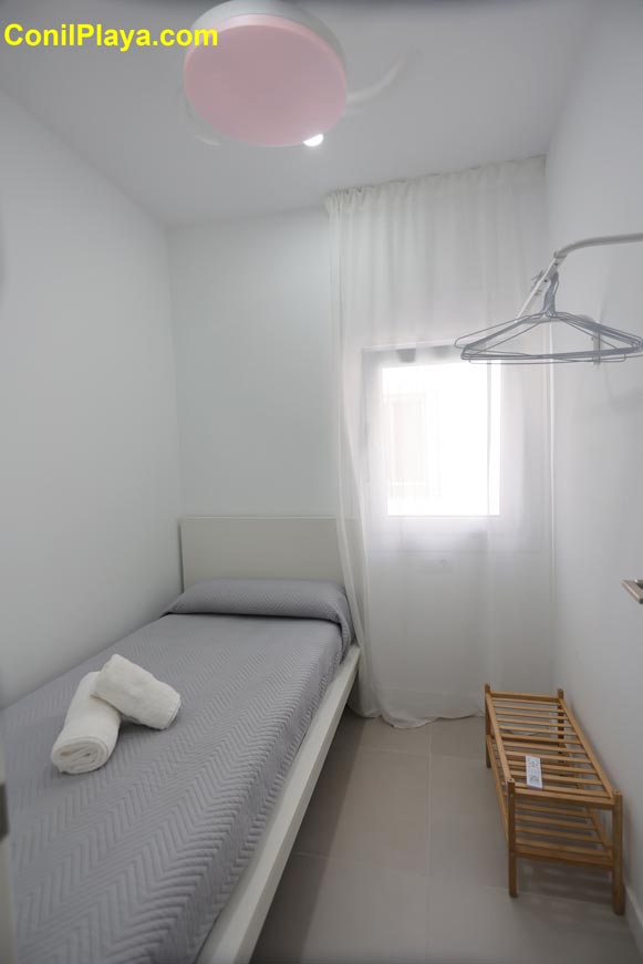 Vista del dormitorio individual blanco con cama, ventana y ventilador de techo y zona para colgar la ropa