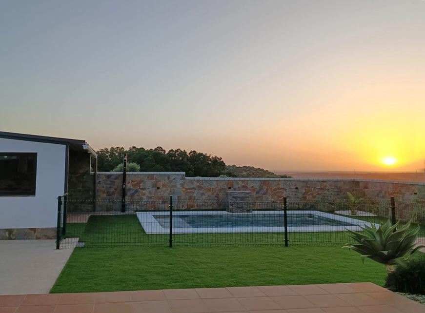 Piscina vallada junto a casa con césped y muro de piedra al atardecer