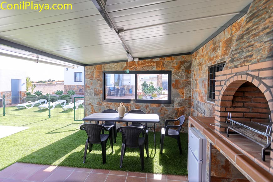 Porche cubierto con barbacoa, mesa y sillas junto a jardín y tumbonas