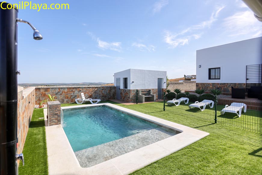 Ducha exterior junto a piscina vallada con césped artificial y tumbonas