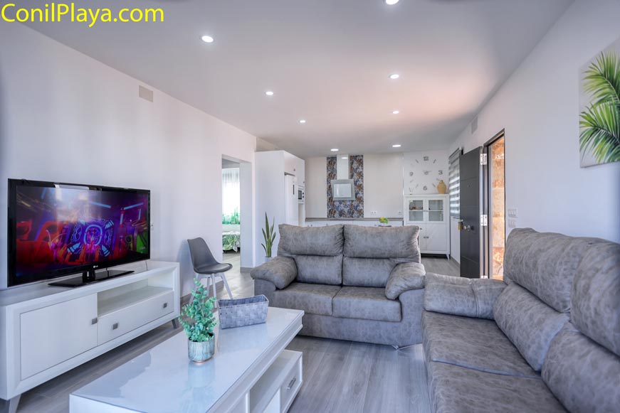 Perspectiva del salón con sofás grises, televisión y mesa de centro blanca, al fondo la cocina
