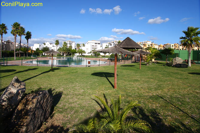 Jardín amplio con piscina, sombrillas de paja, palmeras y edificios residenciales al fondo.