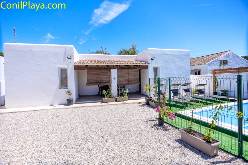 Fachada blanca con porche, piscina vallada y varias macetas en un patio soleado bajo cielo azul