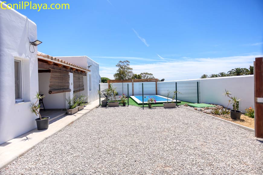 Patio exterior con suelo de grava, piscina vallada al fondo y plantas en macetas junto a pared blanca