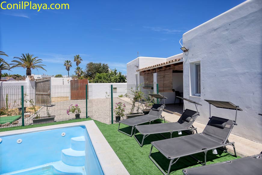 Piscina pequeña junto a varias tumbonas en un patio con césped artificial y edificio blanco bajo cielo azul
