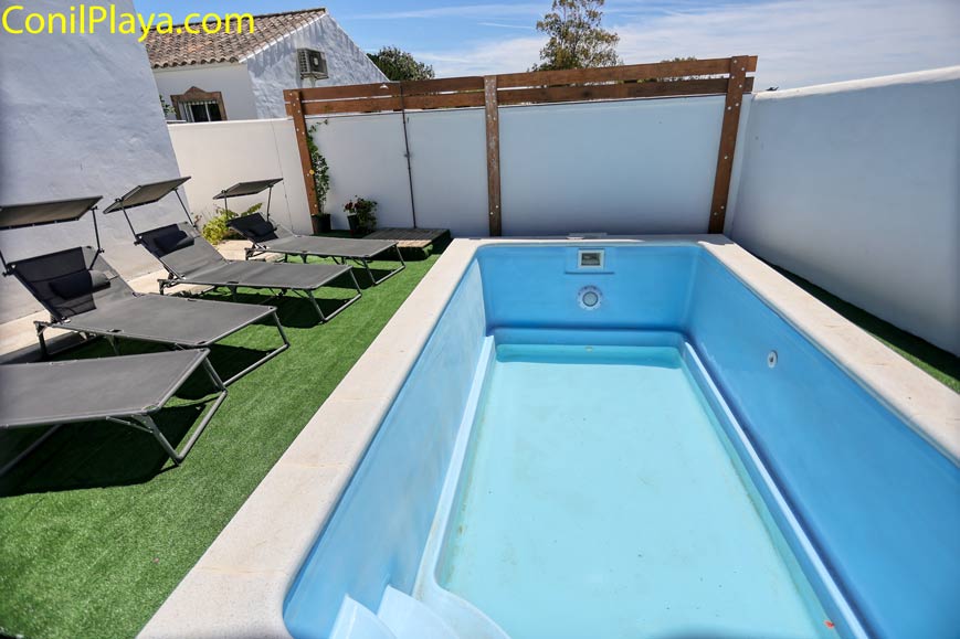 Piscina pequeña rectangular rodeada de césped artificial, hamacas reclinables y paredes blancas.